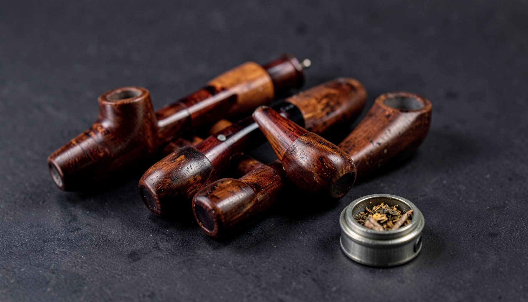 Several polished wooden tobacco pipes resting on a dark surface, with a small tin of aromatic pipe tobacco nearby